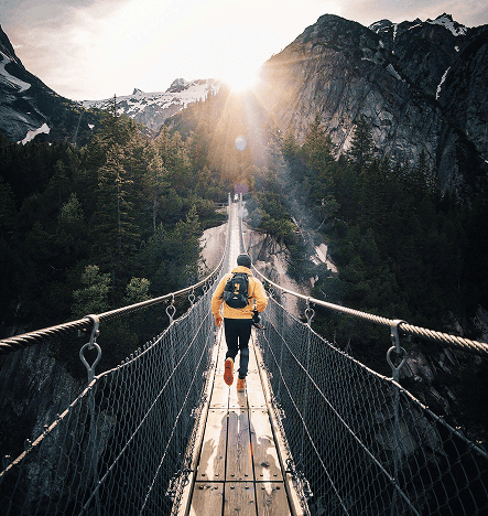 Hiker crossing a suspension bridge in a mountainous landscape at sunrise.
